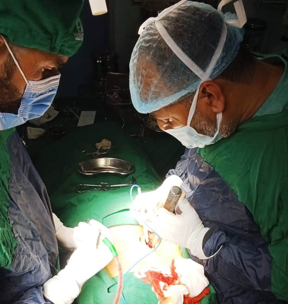 Two surgeons in operating room performing surgery on a patient, wearing green surgical attire and masks.